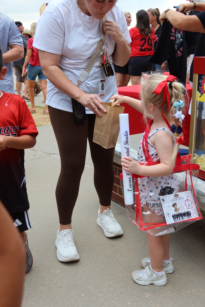 Adult and child interact at an outdoor event, adult hands bag to child, background blurred with people and booths.