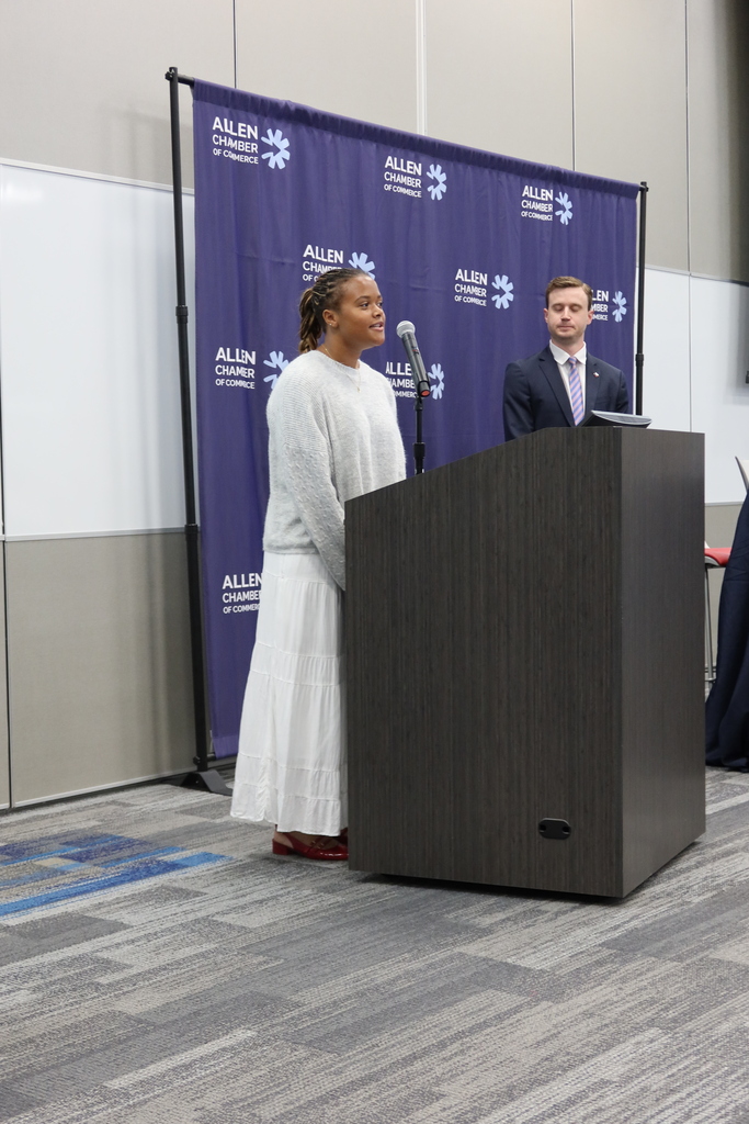 Two individuals, one speaking at a podium and another standing behind it, with a purple banner in the background.