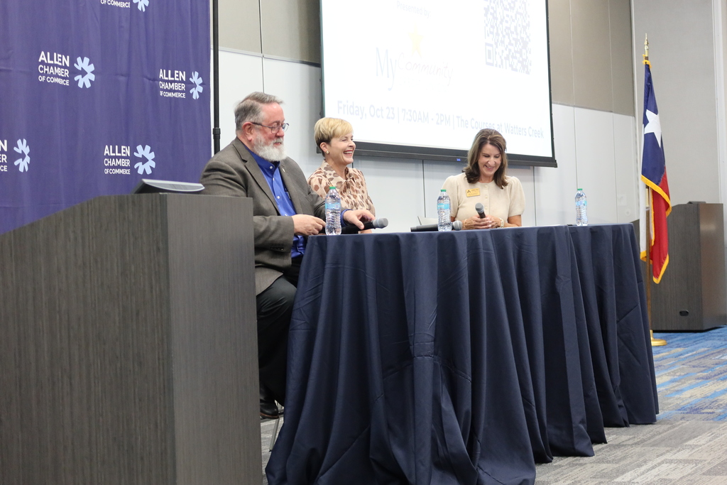 Three people sit at a table with a presentation screen in the background. One person wears a suit, while two others wear blouses.