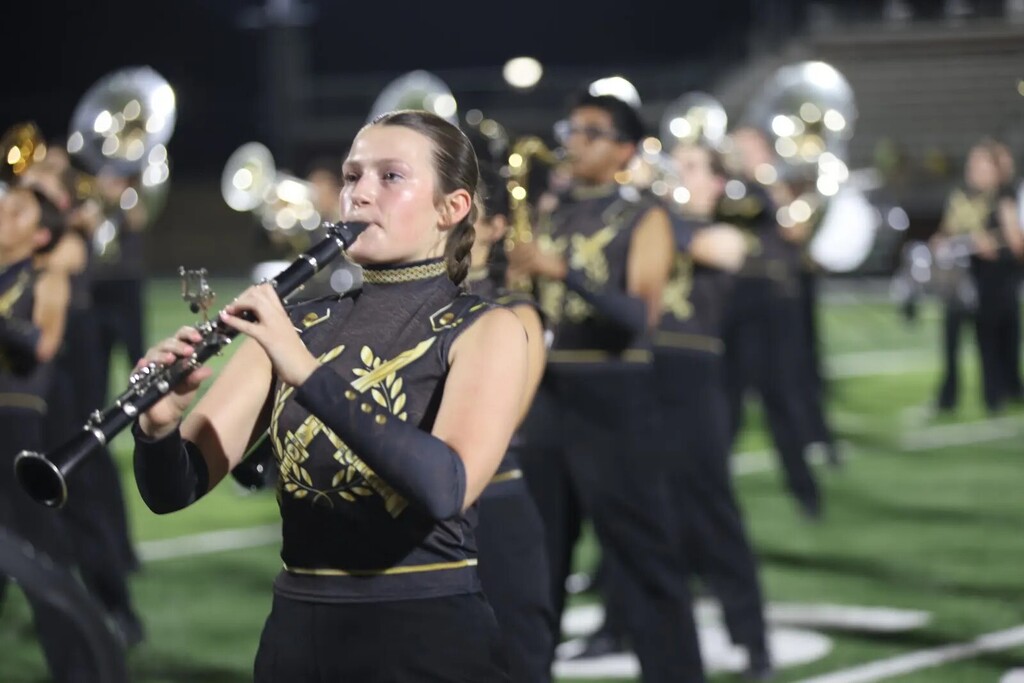 A woman playing a clarinet on a football field with a marching band in black and gold uniforms.