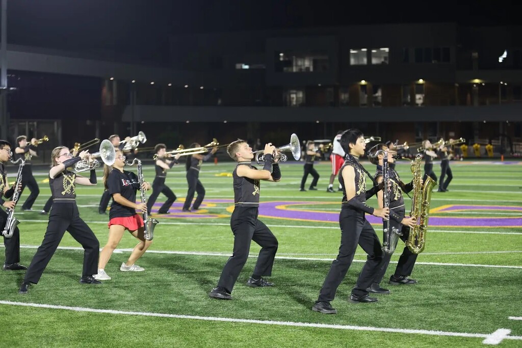 A band performs on a football field at night. Players hold various instruments, including trumpets and saxophones.