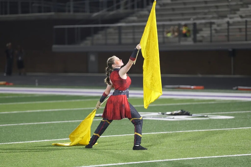 A person in red and black with a yellow flag on a field with empty stands in the distance.