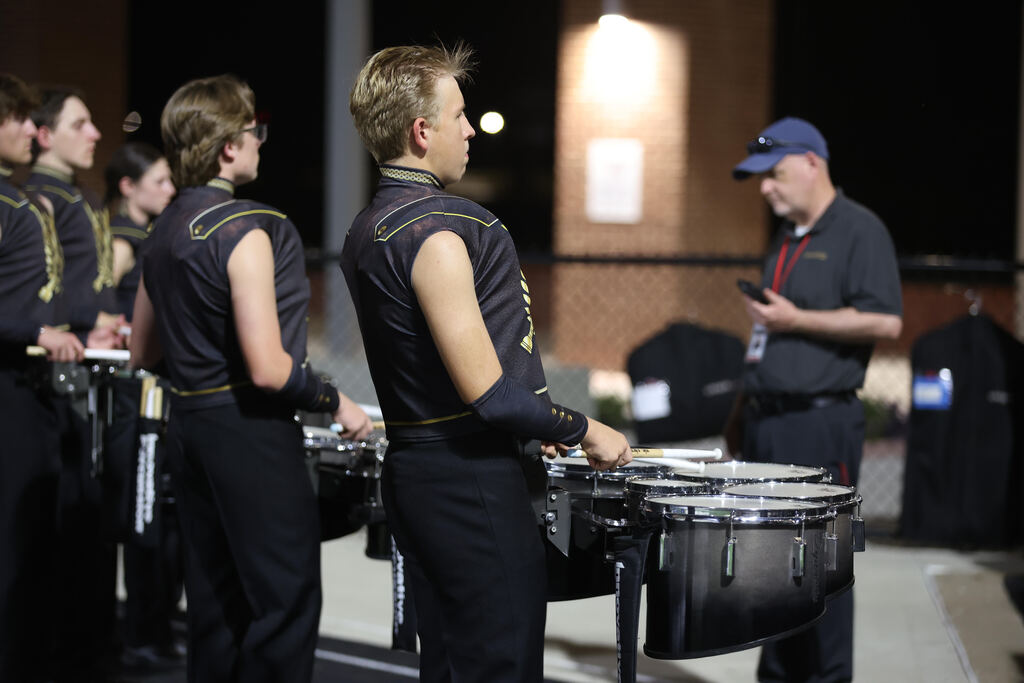 Multiple drummers in uniform perform outdoors at night with a man in a blue hat looking at his phone.