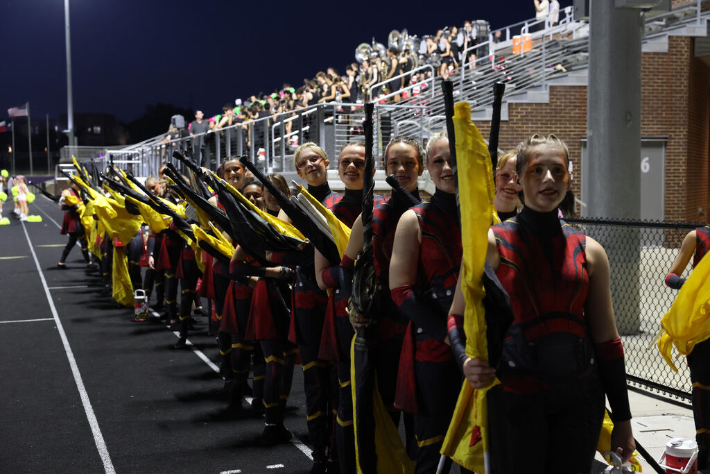 People in red, black, and yellow uniforms line up on the field, holding flags, with bleachers in the background.