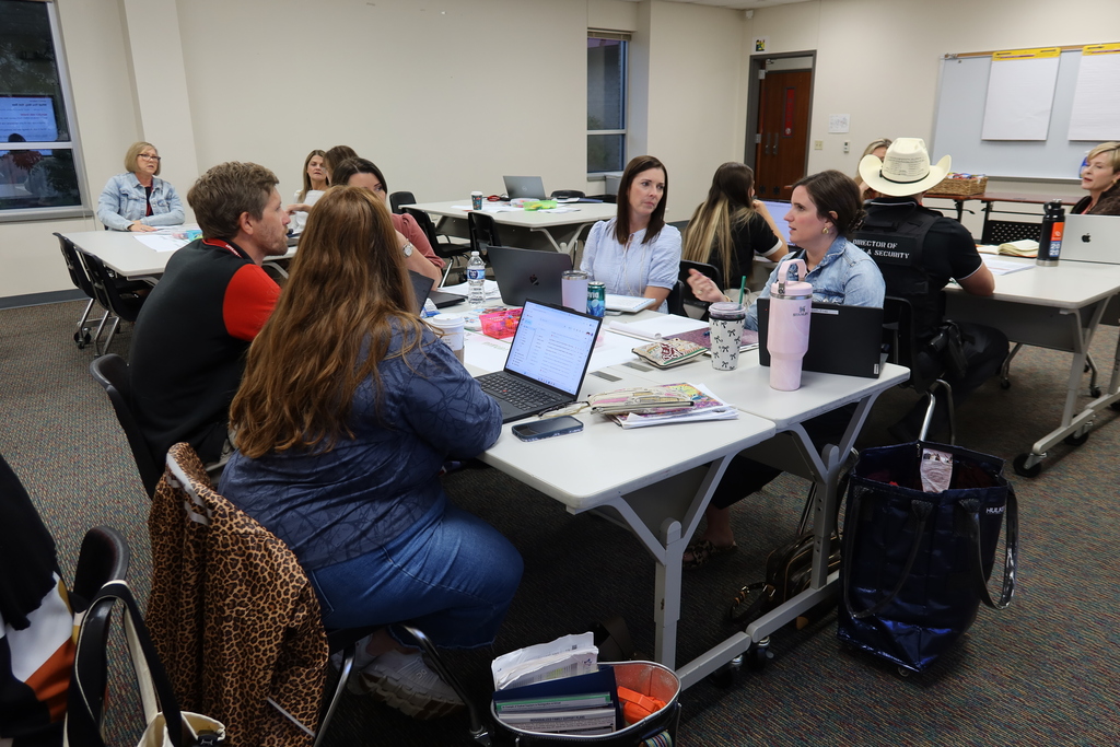 People sit around a table in a room with laptops and books. A woman in a cowboy hat is seated.
