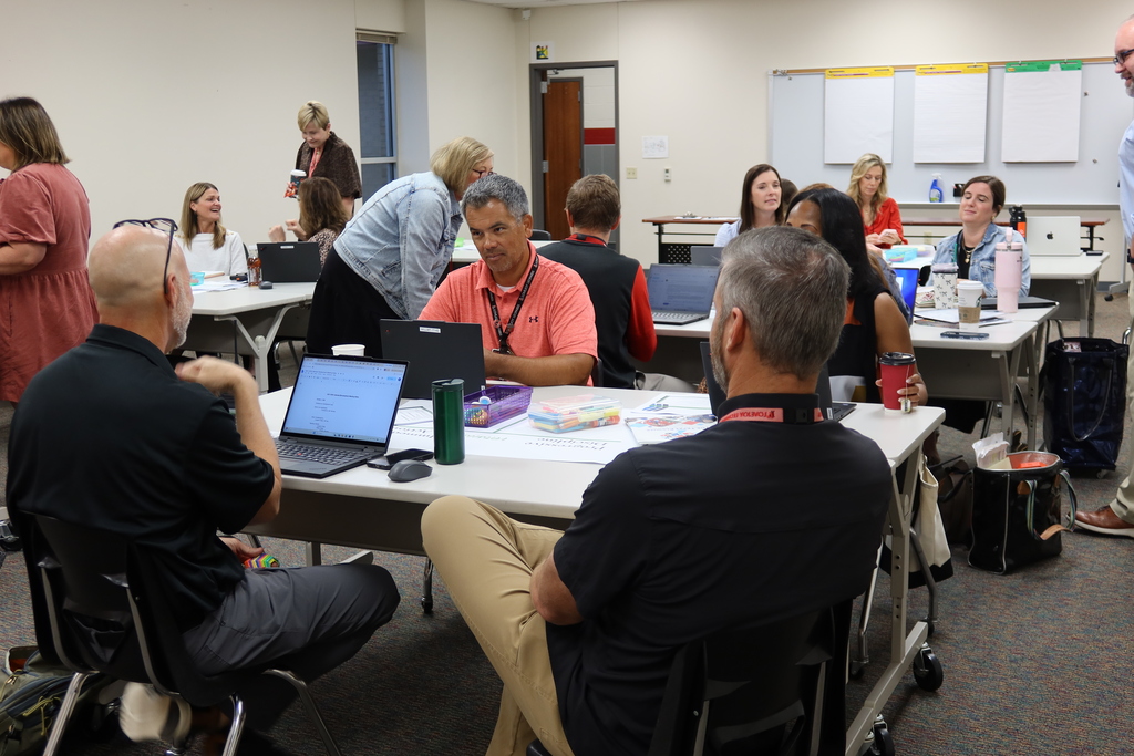 A group of people are seated at tables, working on laptops in a classroom. White boards and tables are visible.