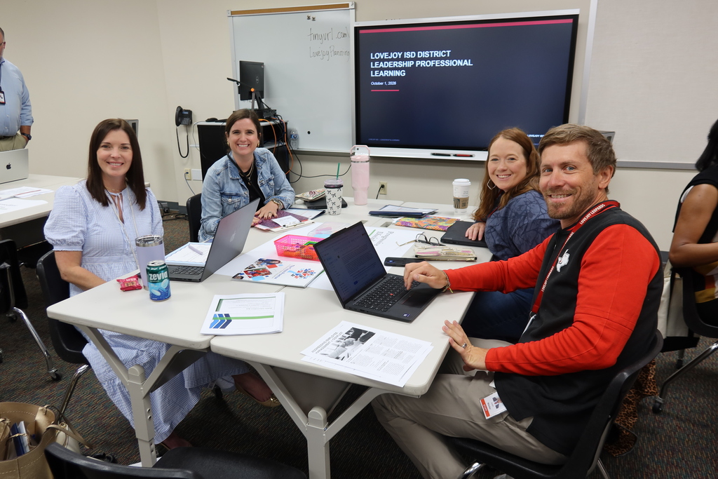 Group of adults sitting around a table with laptops and books. A whiteboard and screen are behind them.