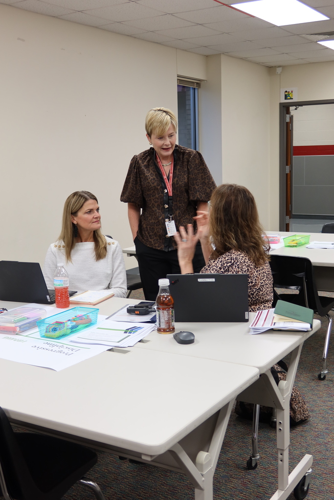 Three women in a room with desks. One woman stands, two sit. Tables have laptops, papers, and bottles.