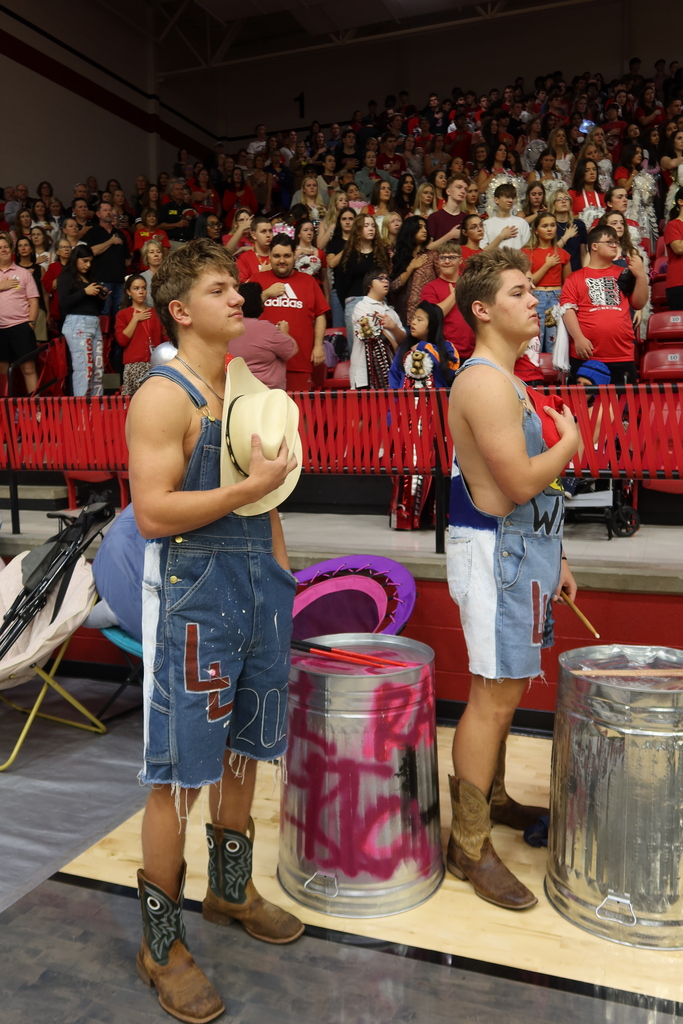 Two men in overalls and cowboy hats standing in front of buckets on a stage with a crowd in the background.