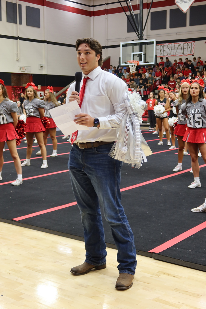 Man standing on basketball court holding paper and microphone, cheerleaders in red skirts and white tops behind him.