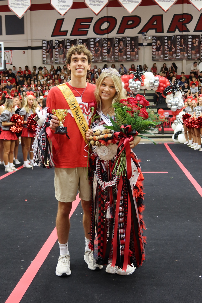 A man and a woman pose with flowers and a trophy on a stage in front of a crowd.