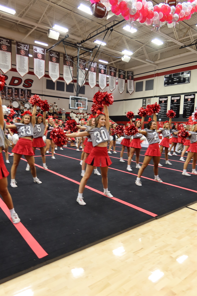A group of cheerleaders perform a routine in a gym, wearing red skirts, white tops, and white shoes.