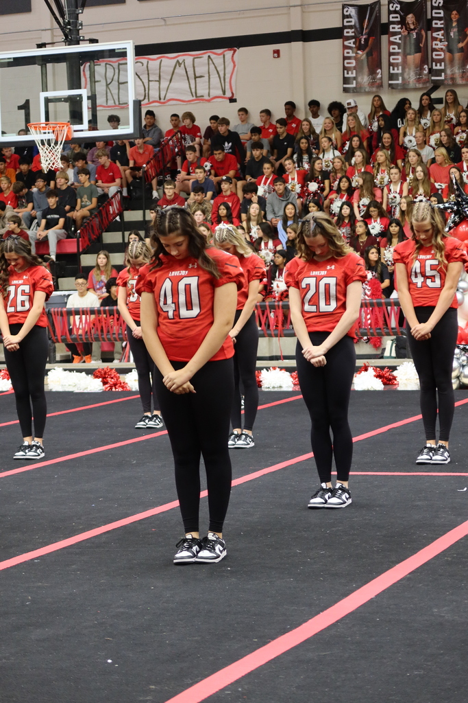 Five cheerleaders in red and black stand on a gym floor with red lines. A crowd in red watches from the bleachers.