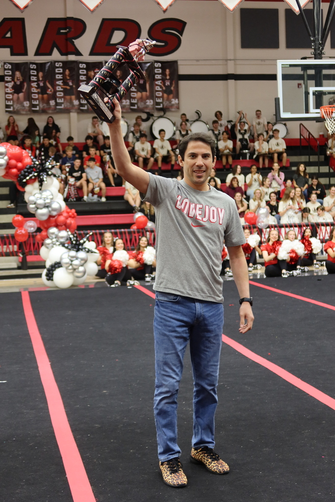 A man in a gray t-shirt and jeans stands on a basketball court, holding a trophy. The audience sits in the background.