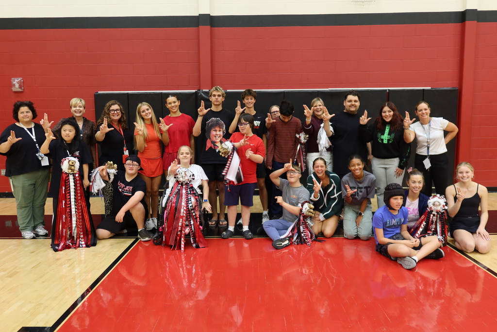 A group of people with pom-poms posing in a gymnasium with red flooring and red walls.