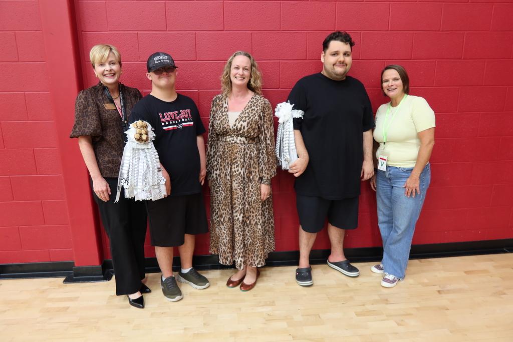 Five people stand in a line in front of a red wall. Each holds a trophy. They wear casual clothing.