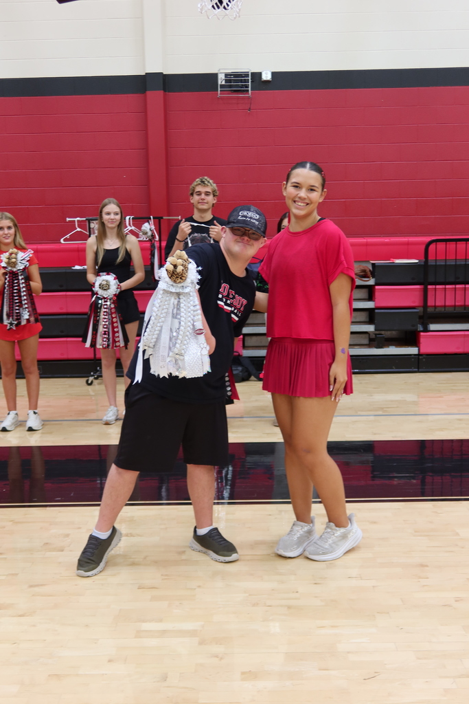 A boy and a girl in a gym. They stand in front of a red wall with bleachers.