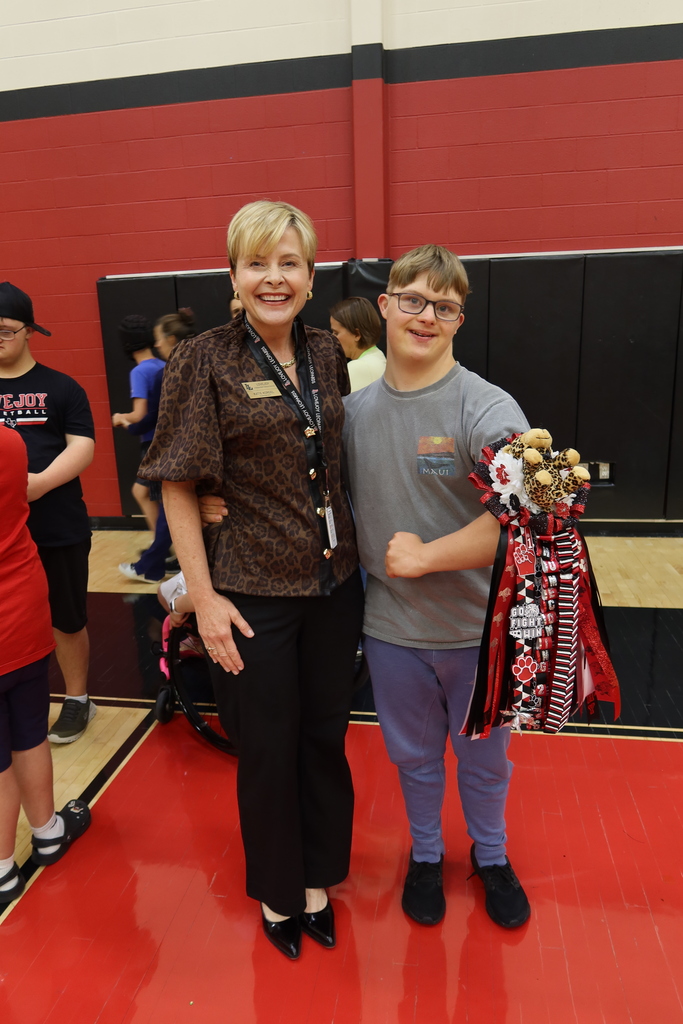Two people stand on a red basketball court with a trophy, near a red wall. A child holds a trophy and a woman stands next to him.