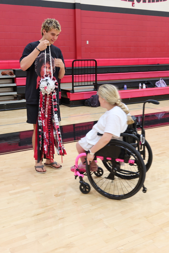 A young person in a wheelchair holds a pom-pom while another individual holds a different pom-pom. Behind them, a red and black wall with bleachers is visible.