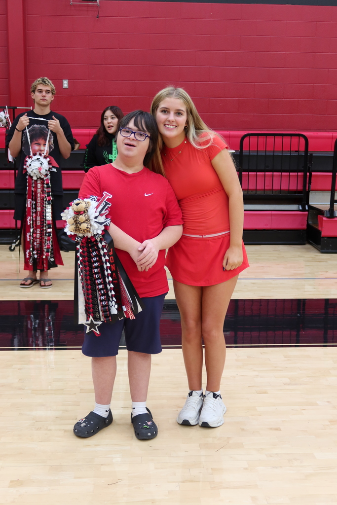 Two people stand on a court, one holding pom-poms. A red wall and bleachers are behind them.