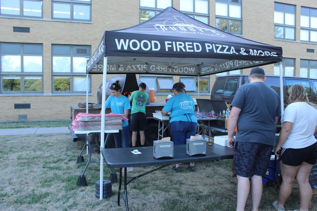 people standing in line for food while staff prepares pizzas