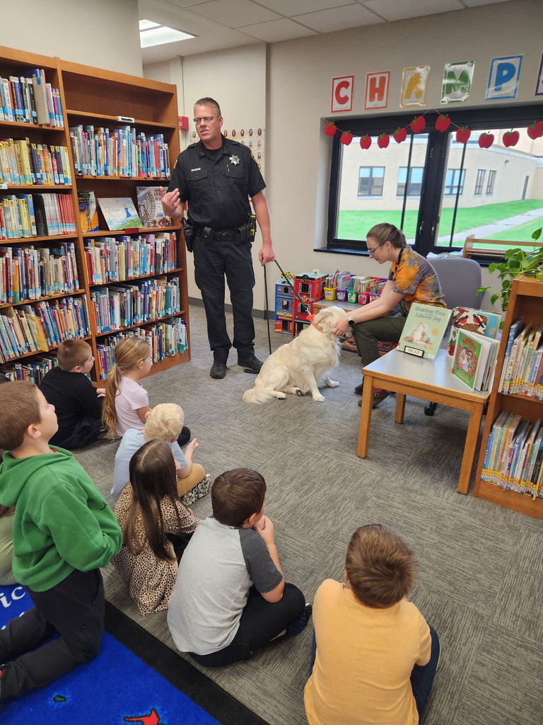 librarian petting dog who is leashed to sheriff deputy and students sitting on the rug watching