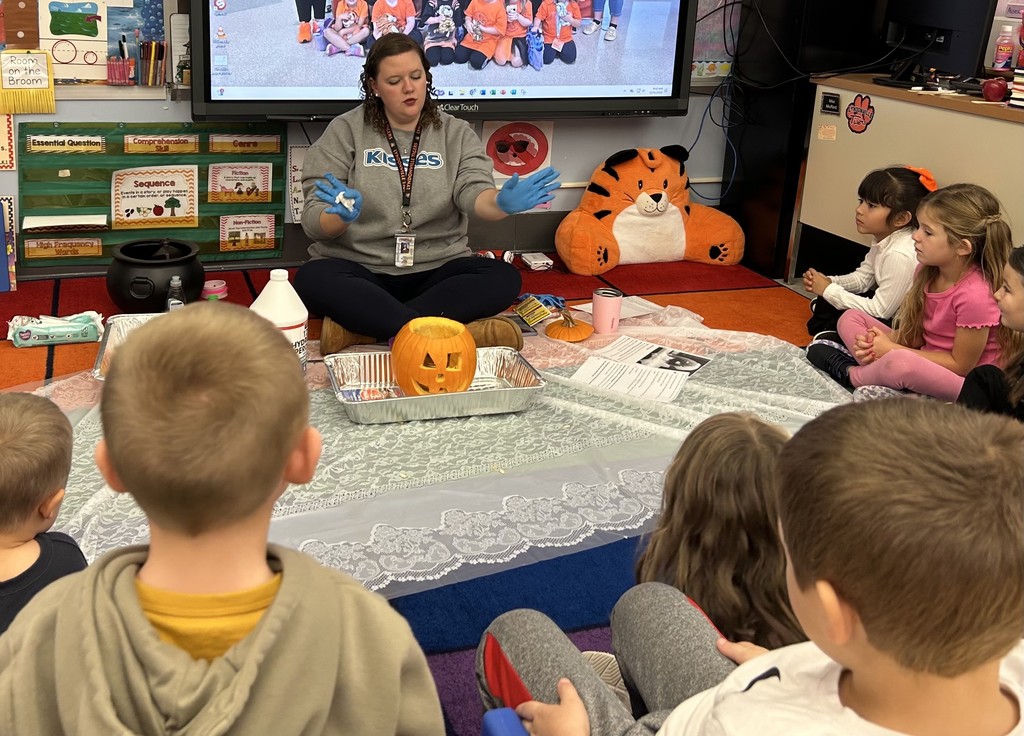 students sitting on rug with their teacher looking at pumpkin experiment