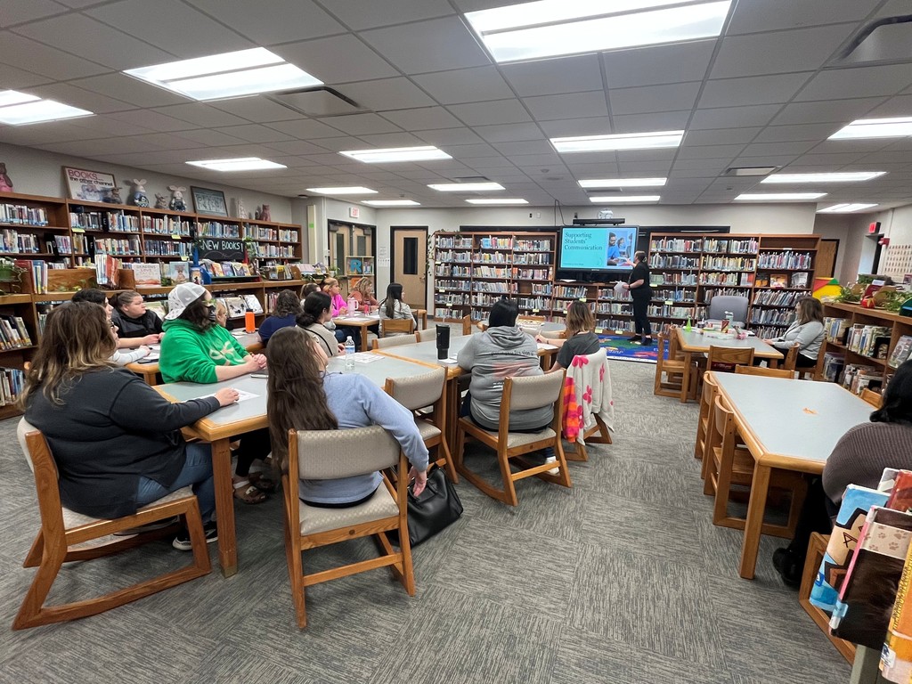 staff sitting at tables in library listening to a speaker in front of a screen with information on it