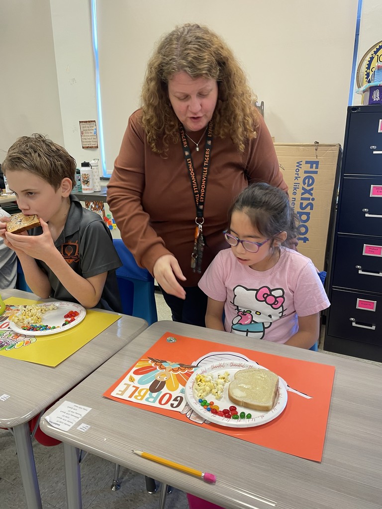 students sitting at table with plates of food and teacher standing nearby