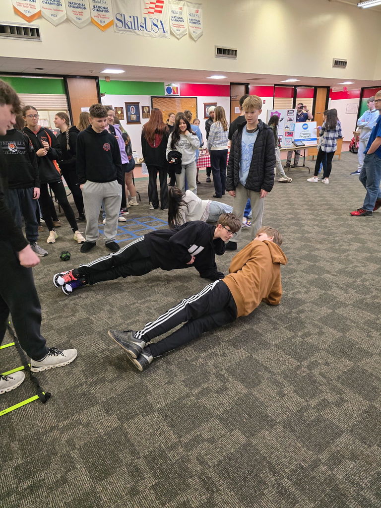 two stufents lying on their sides on the ground while another student instructs them and other students observe them