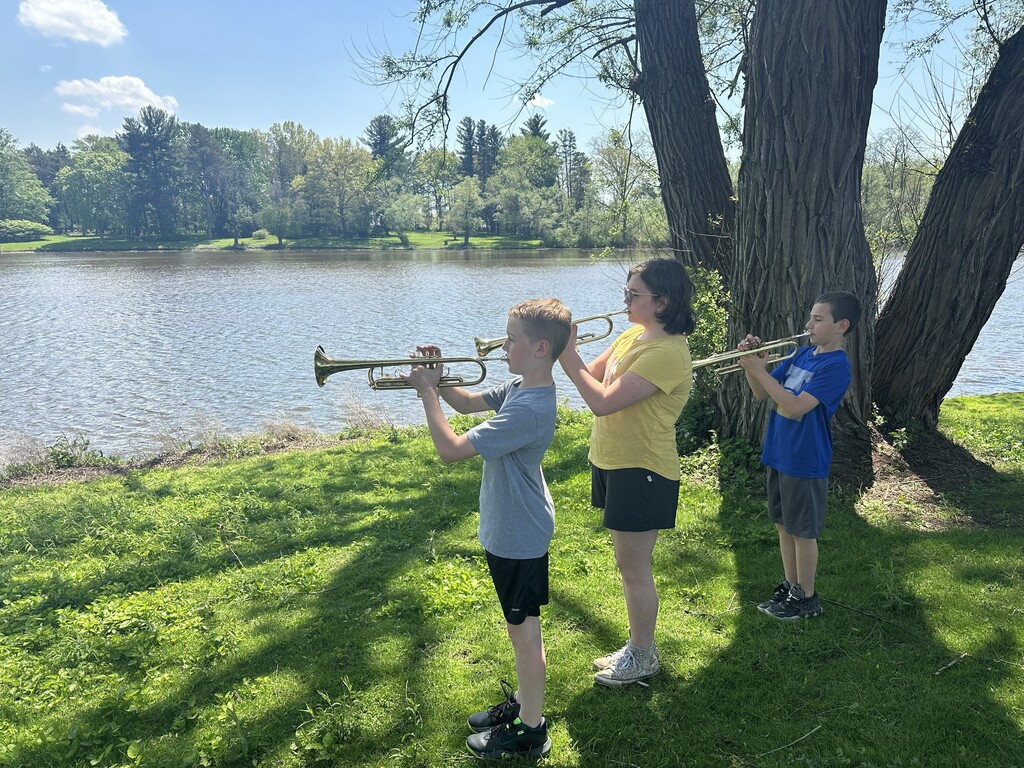three students playing trumpets outside near river