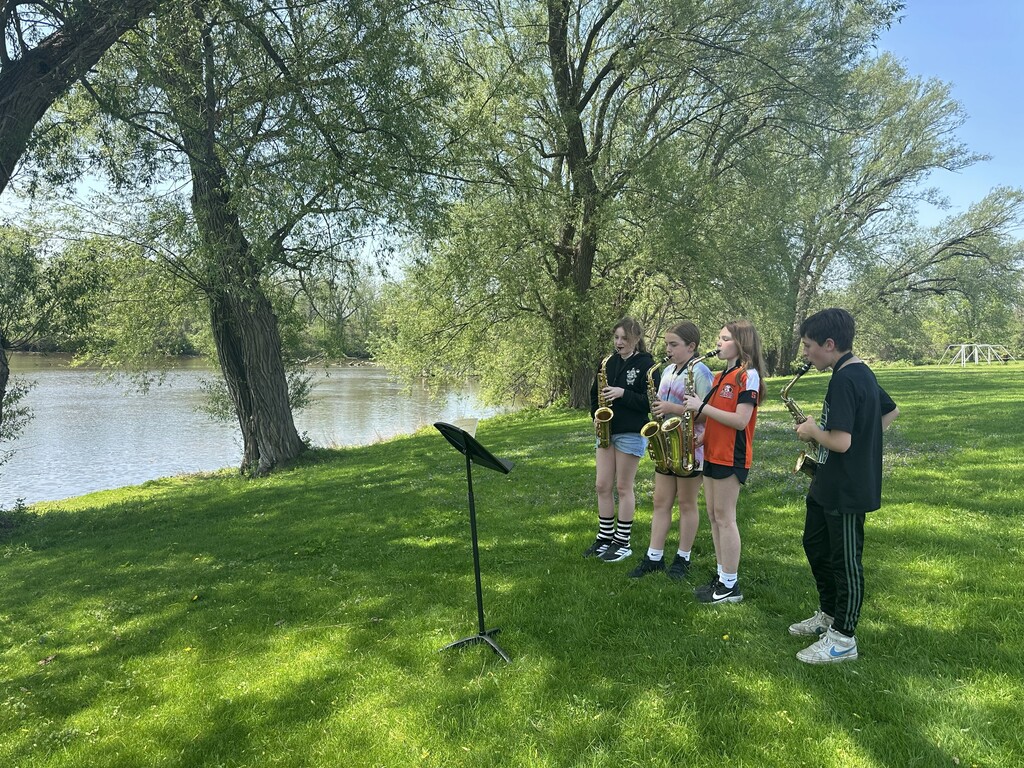 four students playing saxophones outside near river