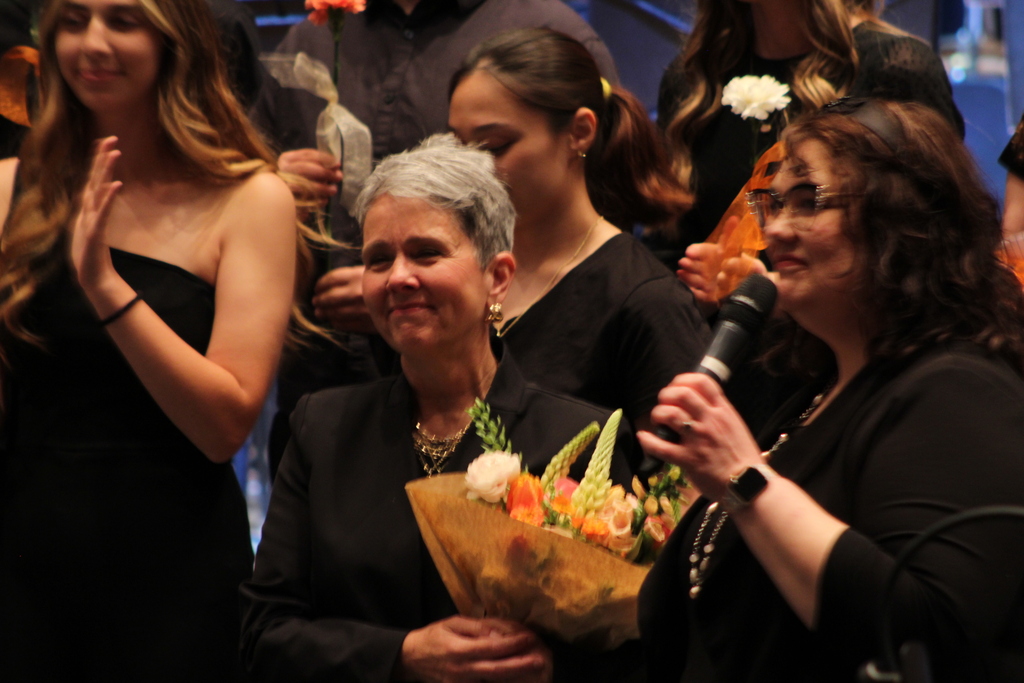 music teacher holding flowers while students clap behind her on risers and fellow music teacher talks into the microphone