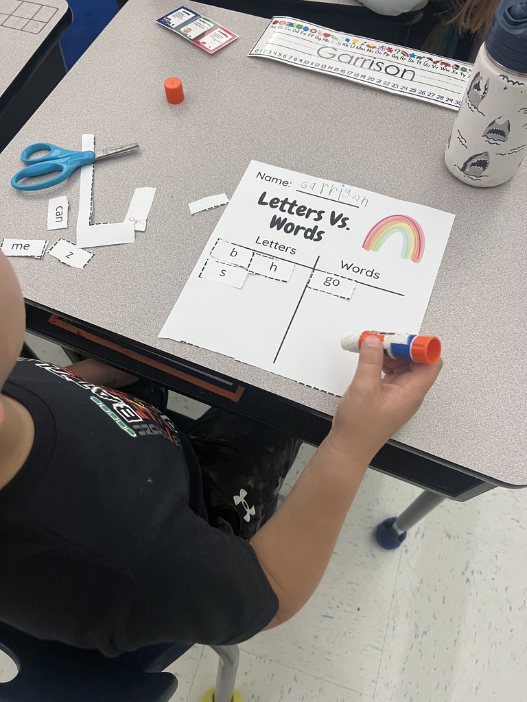 A student sits at a desk using a glue stick to match cutout letters and words on a sheet of paper.