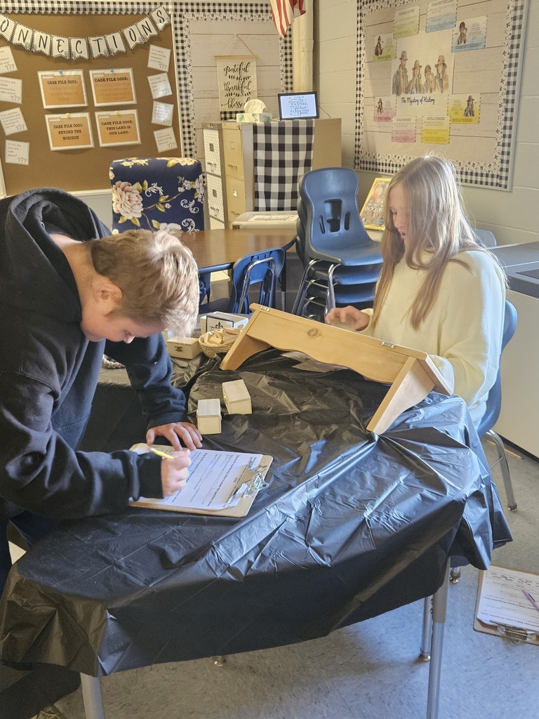 7th Grade Social Studies - learning about the Middle Colonies. A male student takes notes as a female student works on a wodded lap bench.