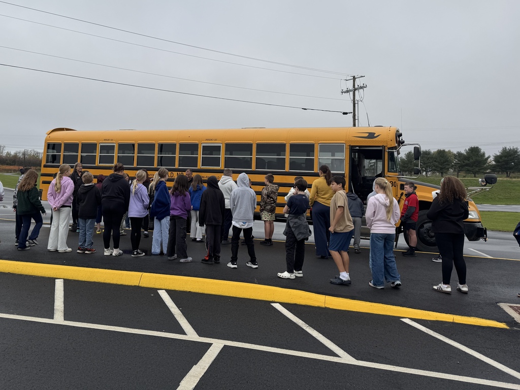 A group of students stand in a line beside a school bus listening to a bus safety talk.