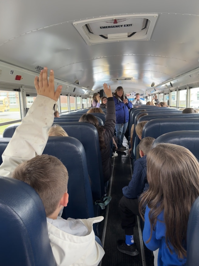 Students seated in a school bus raise their hands in response to a question.