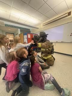 A group of students interact with a firefighter who is kneeling on the floor in full gear
