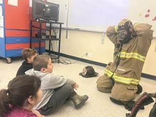 Three students sit on the floor as a kneeling firefighter dons his breathing apparatus