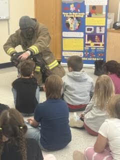 A group of students sit on the floor as a firefighter places his breathing mask on a student's head