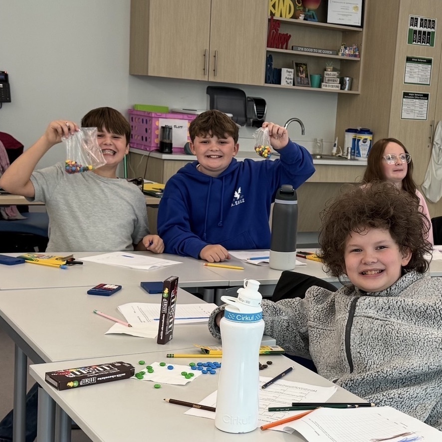 Students sit around a table excitedly holding up the bags of M&Ms they have earned