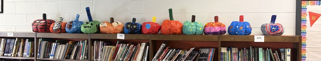 A dozen completed, brightly colored paper mache pumpkins sit atop a bookcase 