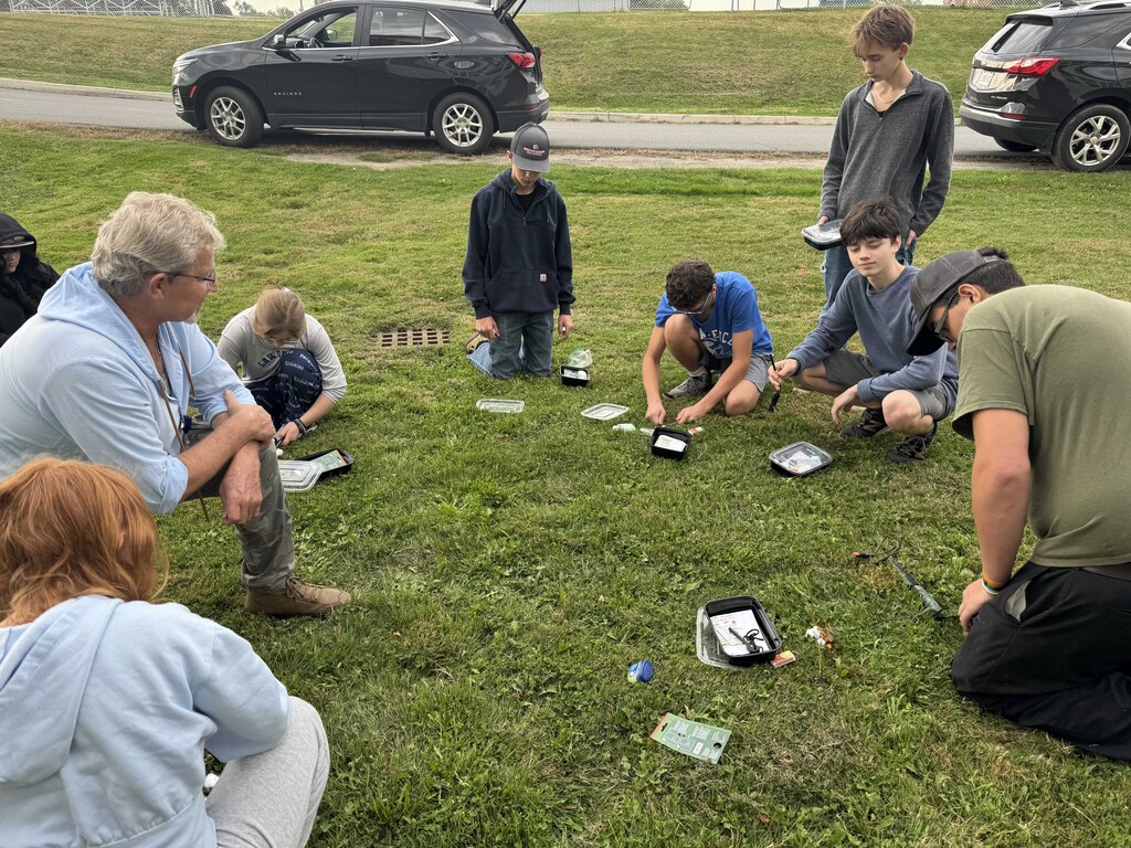 Students are outside sitting in a circle with a teacher learning how to start a fire