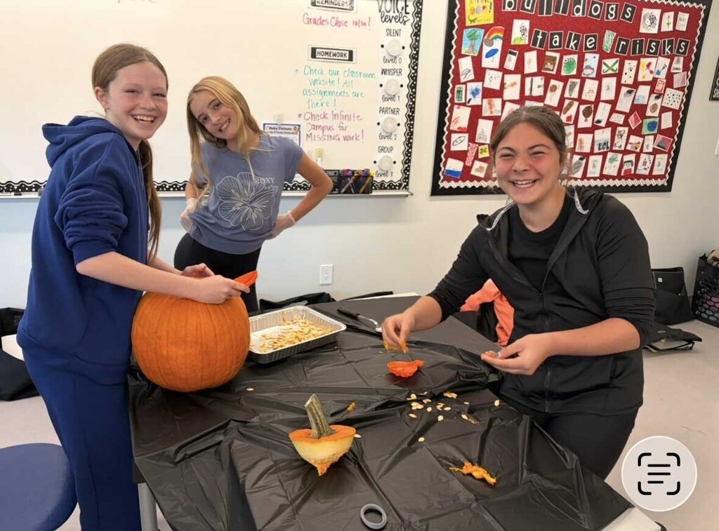 Three girls are carving a pumpkin on a table with seeds and innards in a pan (and all over the table cloth).