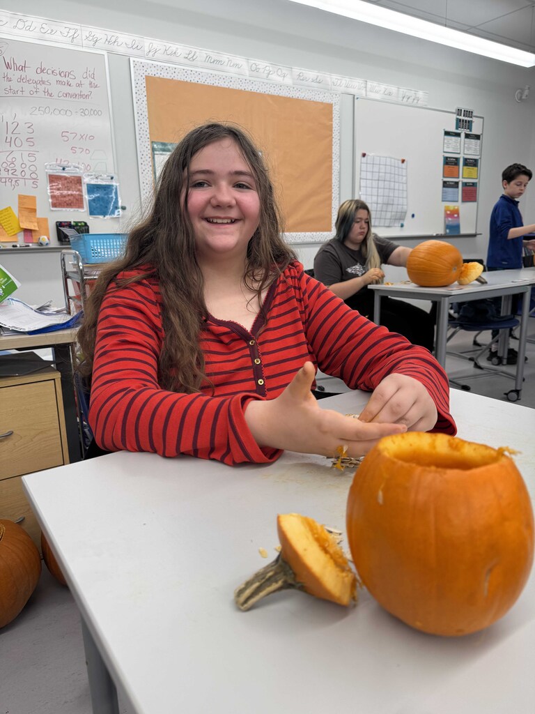 A young lady with long auburn hair sits as she proudly displays her disemboweled pumpkin.