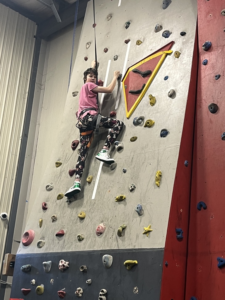 A student, holding a rope and looking at the camera, is half-way up an indoor climbing wall