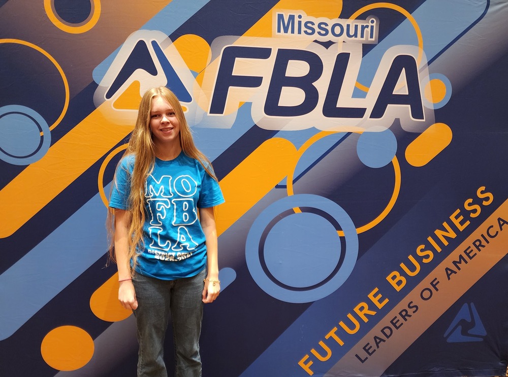 Maddie Moos stands in front of a board reading MO FBLA