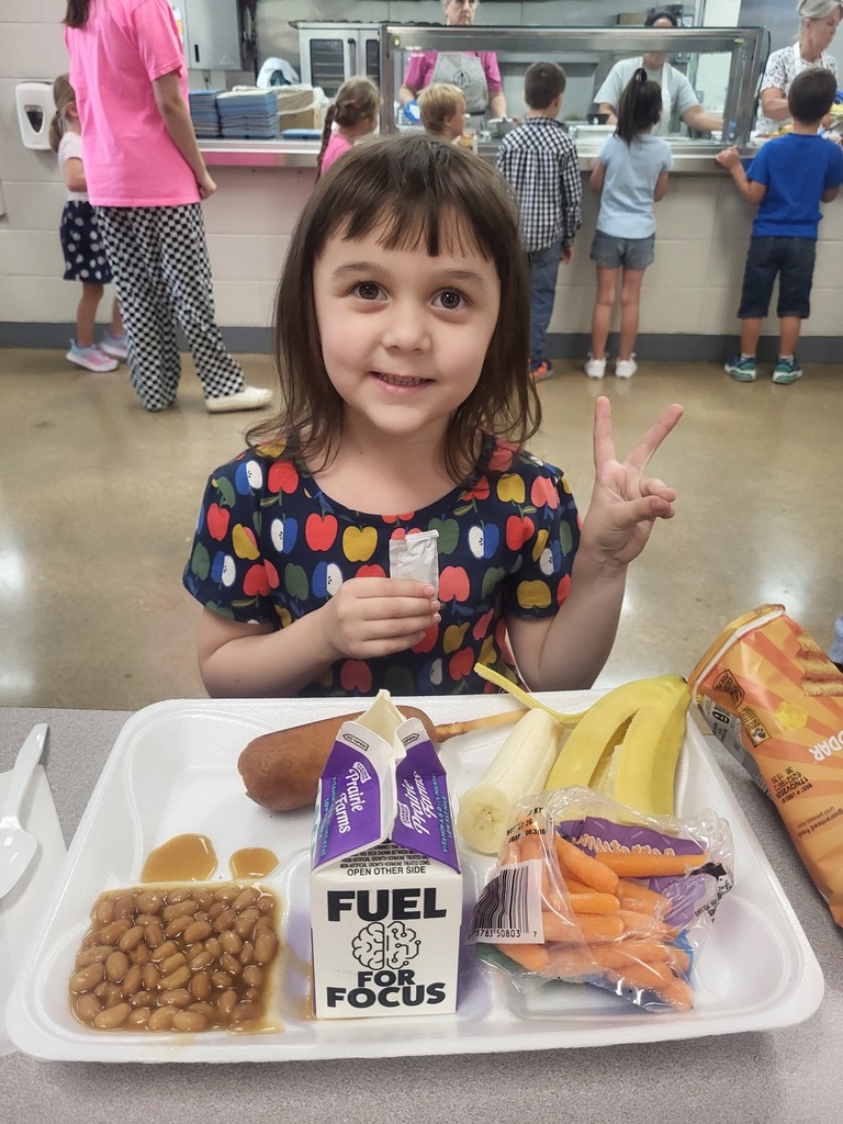 This photo show a little girl eating lunch and smiling for a photo in the cafeteria with other students getting lunch in the background.