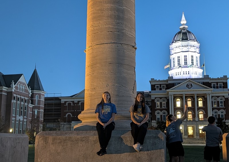 Bailey and Izzy sit next ot a pillar with building behind them lit up in the evening at MU
