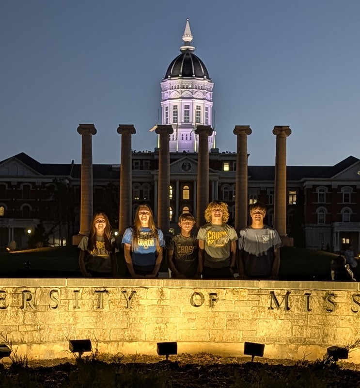 Izzy, Bailey, Parker, Carter and Sebasian are illuminate by a sign in front of the pillars with the tower in the background lit up at night